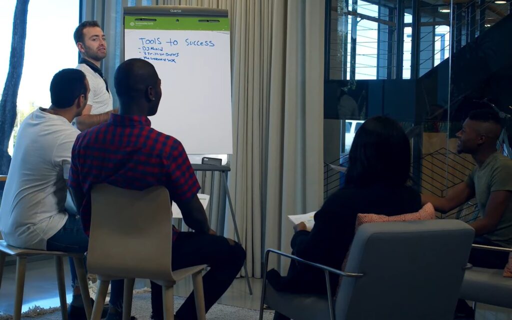 A group engages in a discussion led by a presenter beside a whiteboard